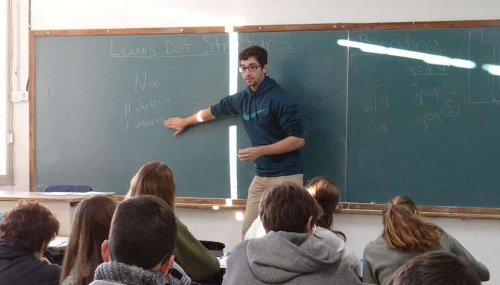 Foto de profesor jóven de gafas enseñando matemáticas con tablero en un salón de clase